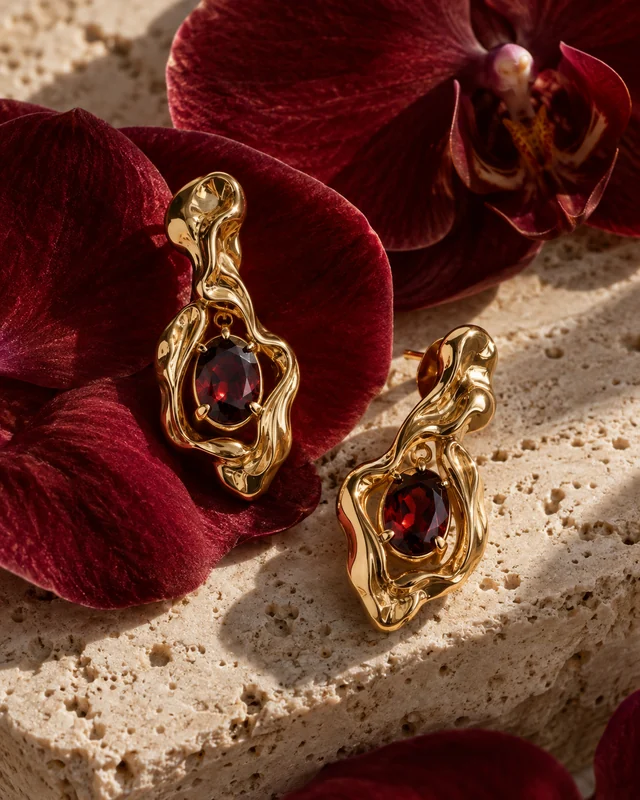 Sculpted gold earrings and garnet among rich red orchid petals and warm ivory travertine.